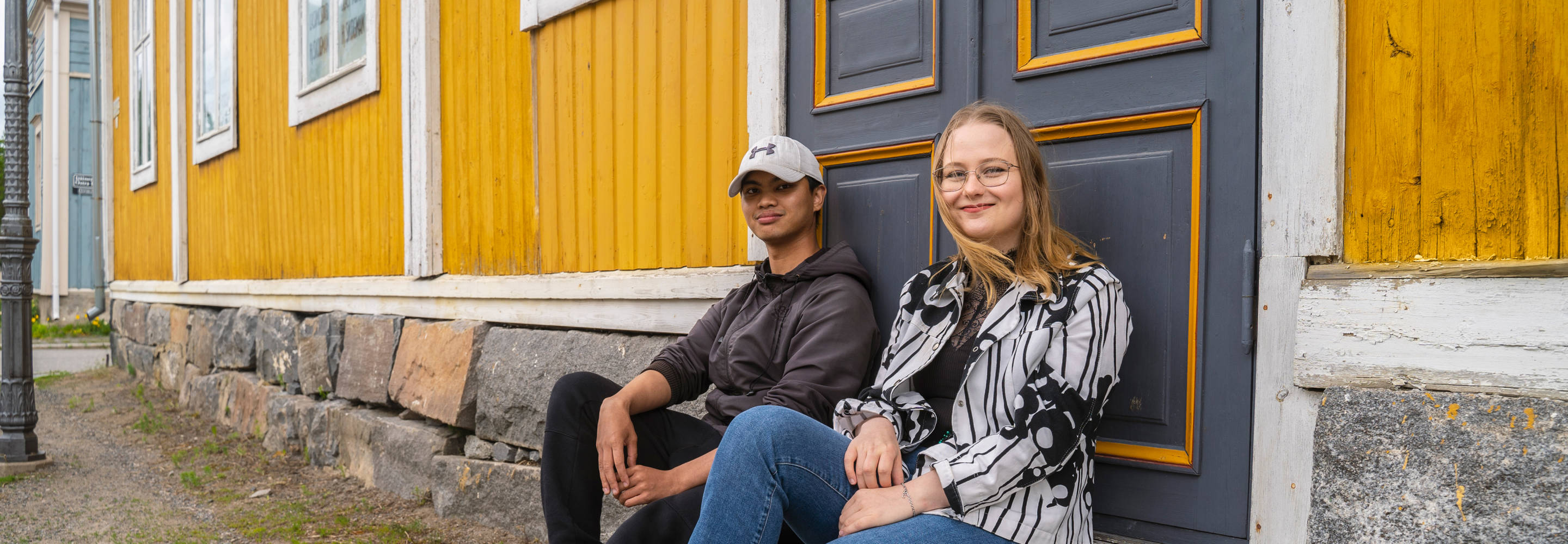 Yhteisöpedagogiopiskelijat Theo ja Sina istuvat Neristanissa vanhan keltaisen talon portailla ja hymyilevät kameralle