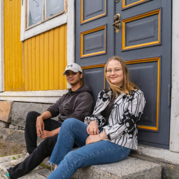 Kaksi yhteisöpedagogiopiskelijaa istuu vanhan talon portailla ja hymyilee kameralle