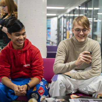 Kaksi opiskelijaa istuu kampuksen käytävällä sohvalla. Toisella on kahvikuppi kädessä ja he nauravat.