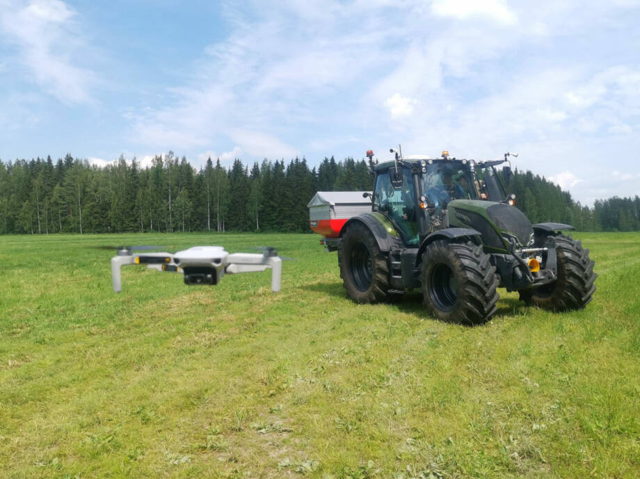 Traktori pellolla kesäpäivänä hiukan kauempana. Kuvan etualalla lentää drone. 